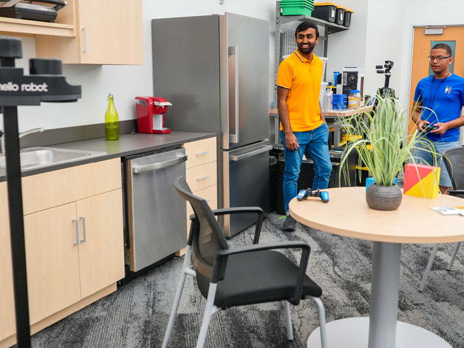 two men in a kitchen environment testing a robot on the other side of the room